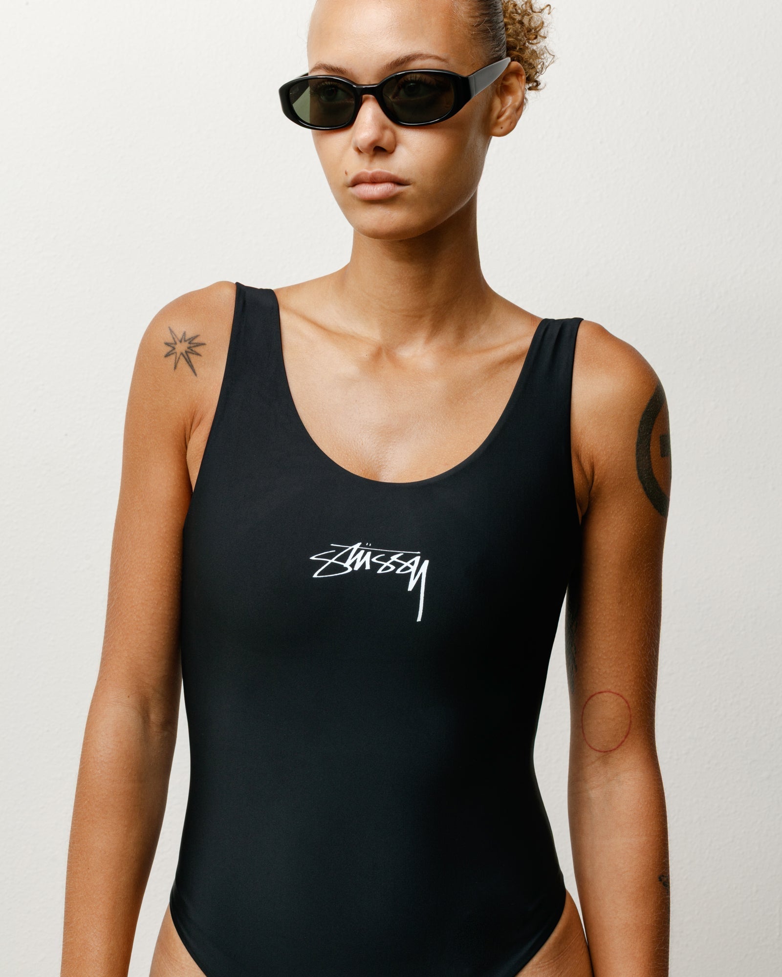 Stüssy 서프 스톡 원피스 수영복 블랙 색상을 착용하고 가슴 부분의 화이트 로고가 돋보이는 모델의 정면 모습.