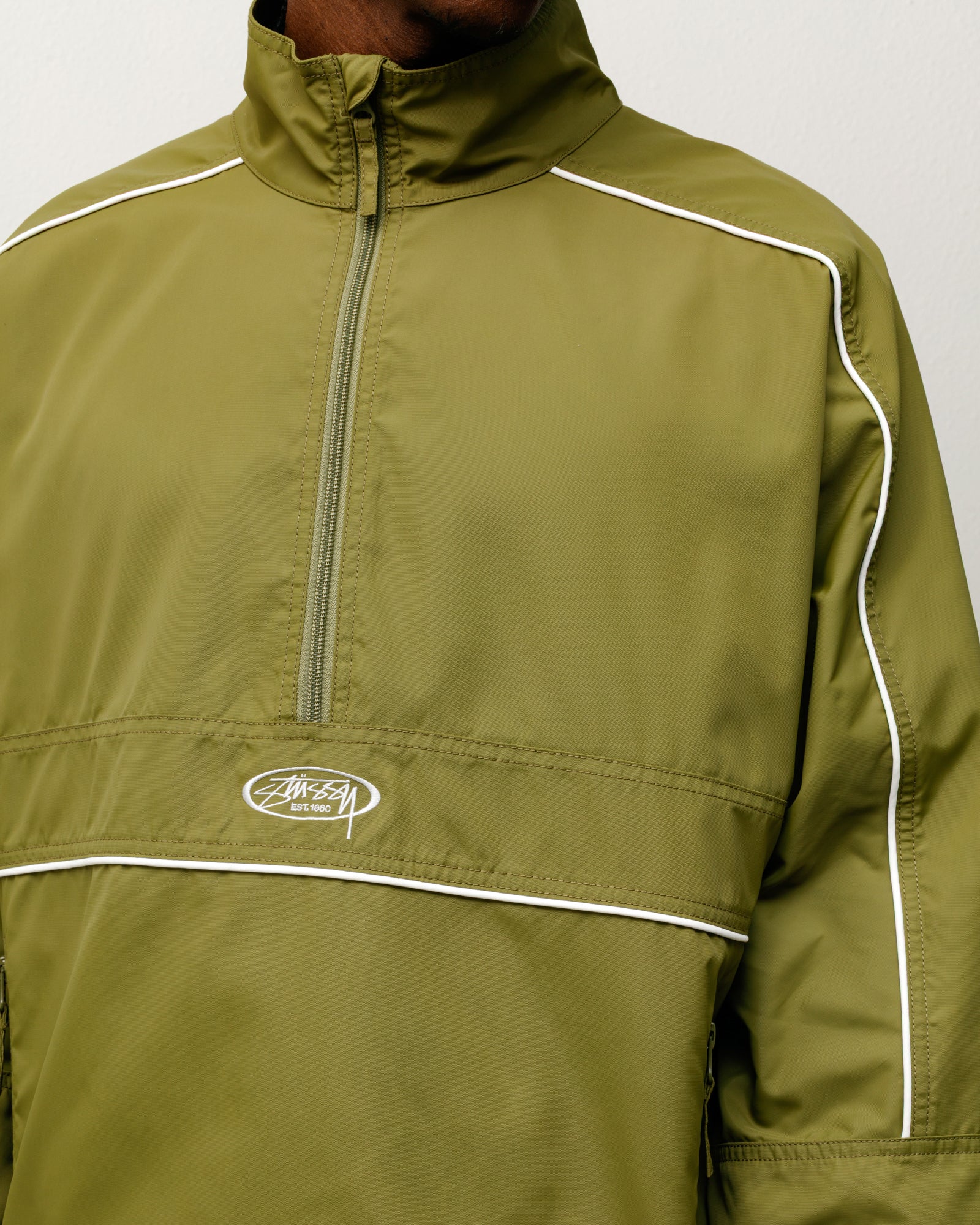 올리브 컬러의 남성용 롱 슬리브 라이트웨이트 풀오버 재킷으로, 가슴의 Stüssy 로고 자수와 화이트 파이핑 디테일이 돋보이는 가벼운 소재의 아우터입니다.