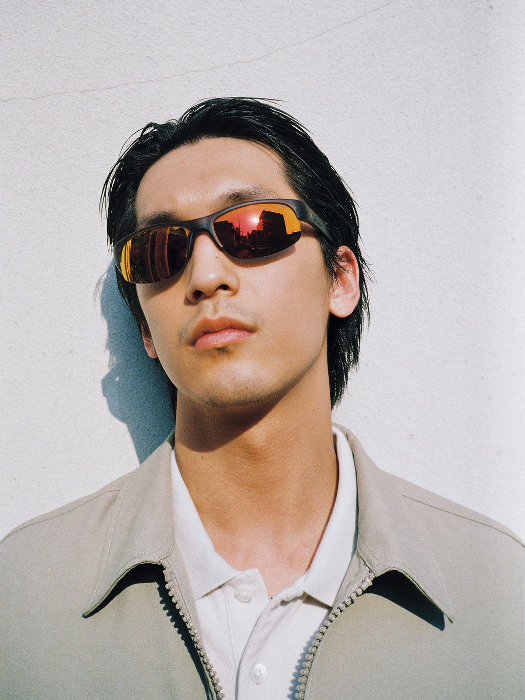 Model leaning against a wall wearing the Hiro Sunglasses in Frost Brown/Red Mirror Lens and Mechanic Jacket in Khaki.