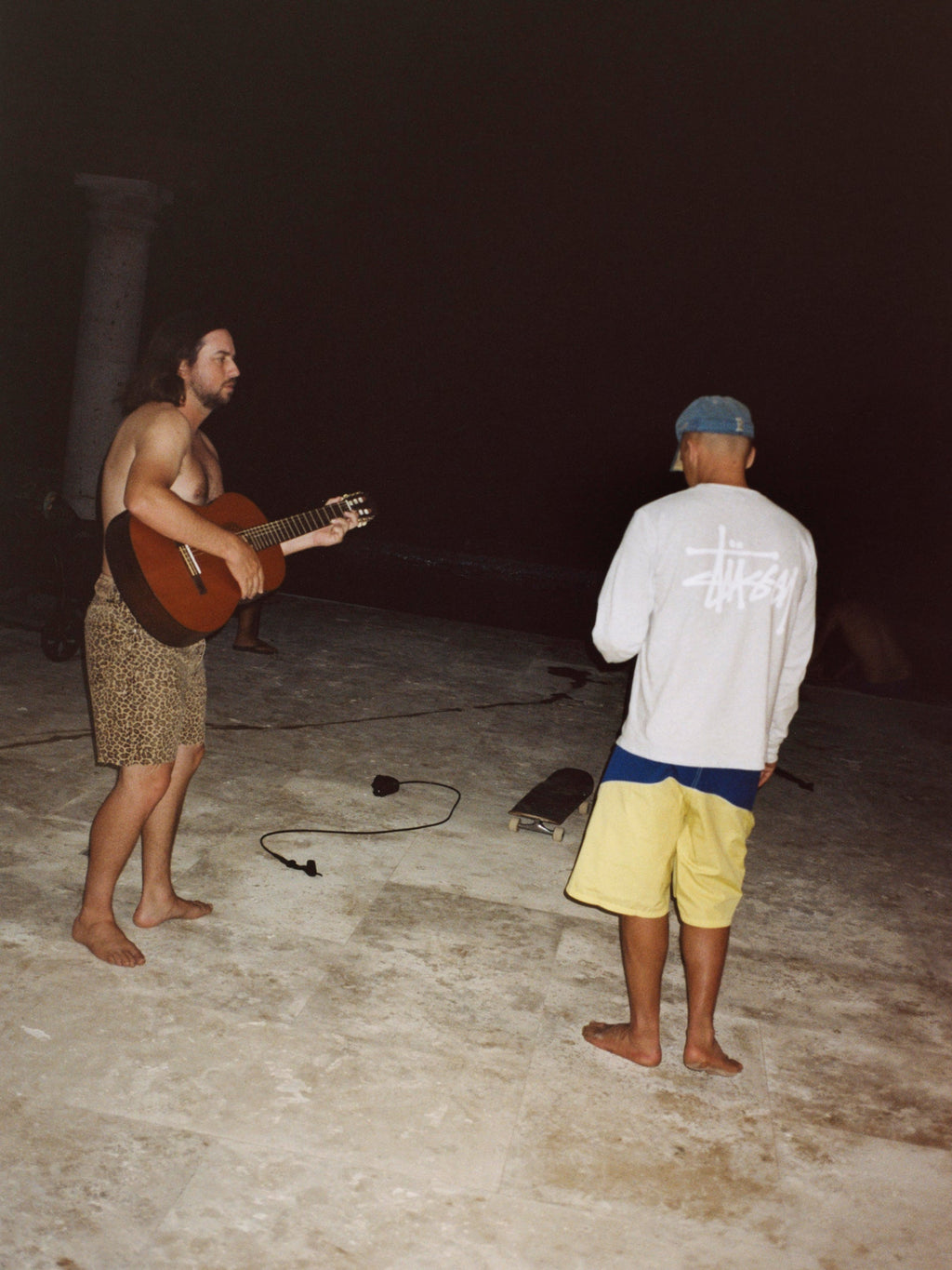 A shirtless man plays an acoustic guitar at night for a friend wearing the Basic Stüssy LS Tee in Ash Heather and Cove Board Short in Yellow.