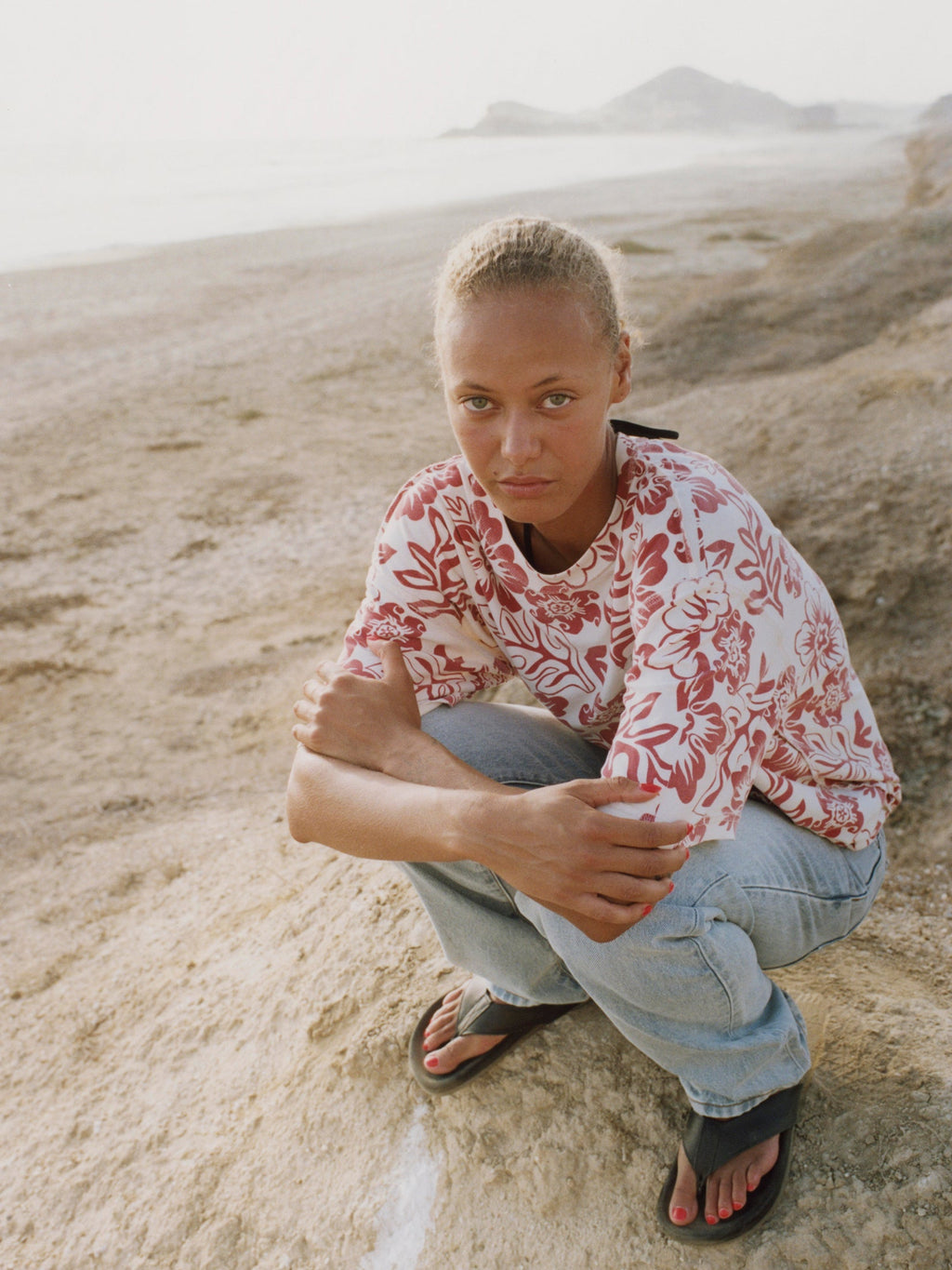 A model wearing the Lazy Floral Tee in White and light wash jeans crouches on a sandy slope.