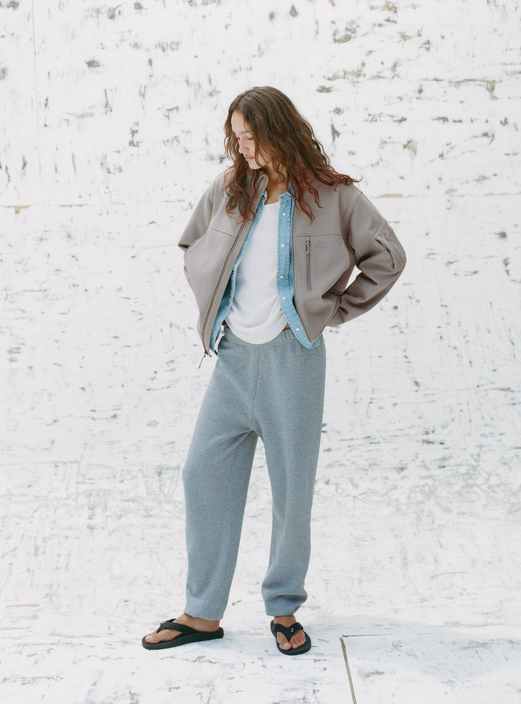 A model stands against a textured white backdrop wearing the Soft Shell Jacket in Moss Grey, Denim Western Shirt in Light Denim, and Beach Slippers in Black.